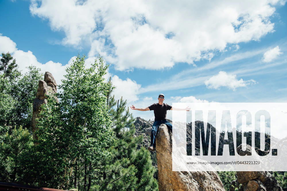 Man with arms outstretched sitting on rocks in sunny mountain scene Keystone, South Dakota, United States CR_KIFR240402-1377296-01 ,model released, Symbolfoto
