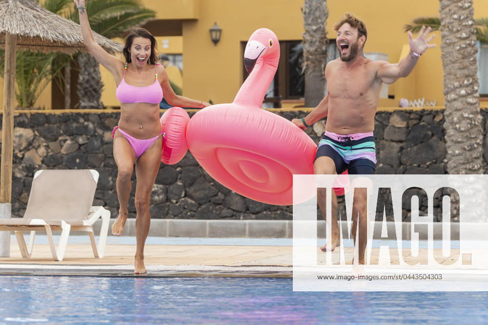 An ecstatic couple is captured in mid-motion, joyfully playing with a giant pink inflatable flamingo by a sunlit swimming pool Model Released JuanAizpuru_ID20330_601362_015