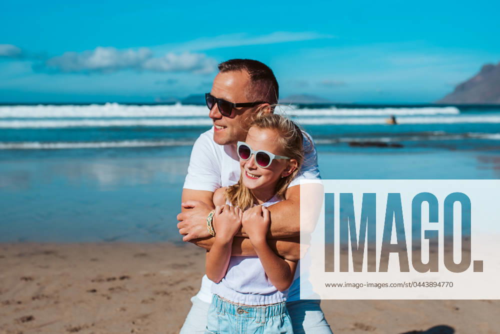 Father embracing daughter on beach. Dad and girl enjoying sandy beach, looking at crystalline sea in Canary Islands. Concept of beach summer vacation with kids. Father embracing daughter, standing on beach. Dad and girl enjoying sandy beach, looking at crystalline sea in Canary Islands. Concept of beach summer vacation with kids. model released