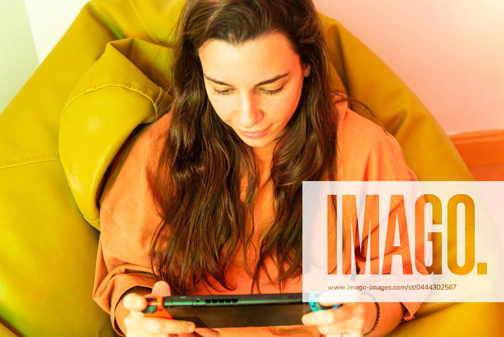 IMAGO Creative, Lifestyle, Gaming A focused gamer sits comfortably, deeply immersed in playing a game on a handheld console, with a vibrant yellow chair adding a pop of color Model Released NataliaPenado_ID20537_606564_001
