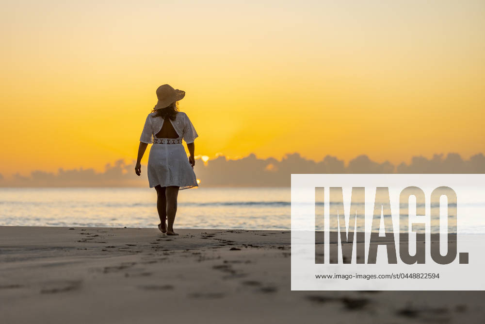 Back view of an unrecognizable woman enjoying a peaceful walk along the shore at sunrise, with the warm glow of the sun casting a tranquil backdrop. Model Released JuanAizpuru_ID20865_615527_019