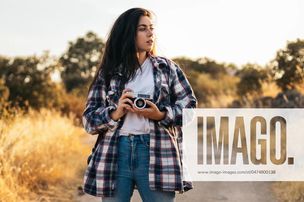 Young woman in checkered shirt and casual clothes taking photos with an analog camera in the field at sunset. The image conveys the concept of retro and vintage Model Released PedroMerino_ID19010_565591_011