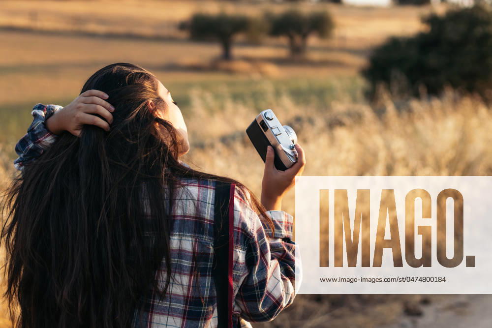 Young woman in checkered shirt and casual clothes taking photos with an analog camera in the field at sunset. The image conveys the concept of retro and vintage Model Released PedroMerino_ID19010_565592_012