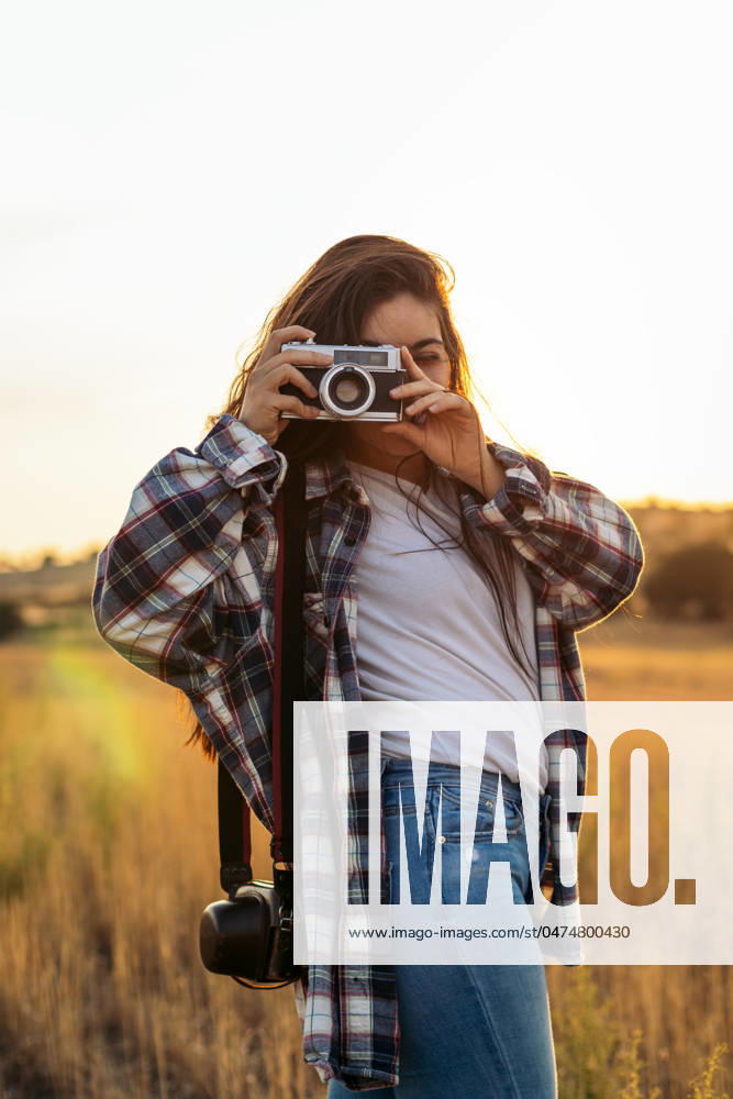 Young woman in checkered shirt and casual clothes taking photos with an analog camera in the field at sunset. The image conveys the concept of retro and vintage Model Released PedroMerino_ID19010_565598_016