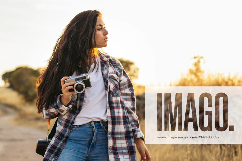 Young woman in checkered shirt and casual clothes taking photos with an analog camera in the field at sunset. The image conveys the concept of retro and vintage Model Released PedroMerino_ID19010_565605_021