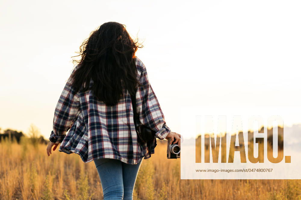 Young woman in checkered shirt and casual clothes taking photos with an analog camera in the field at sunset. The image conveys the concept of retro and vintage Model Released PedroMerino_ID19010_565607_023