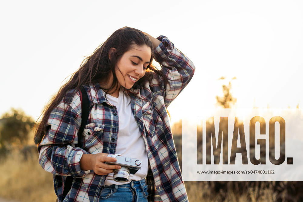 Young woman in checkered shirt and casual clothes taking photos with an analog camera in the field at sunset. The image conveys the concept of retro and vintage Model Released PedroMerino_ID19010_565619_029