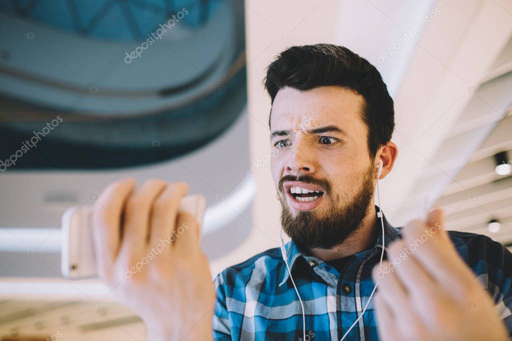 Portrait of a young handsome man with phone and headphones in urban background. Guy is embarassed, his phone stopped working and he doesnt know why. He is looking at mobile and trying to figure out model released