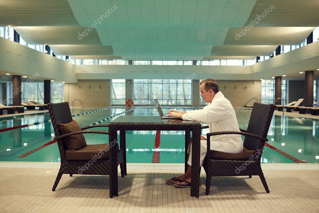 Full length side view of successful senior man using laptop while relaxing at luxury SPA resort with swimming pool in background, copy space model released