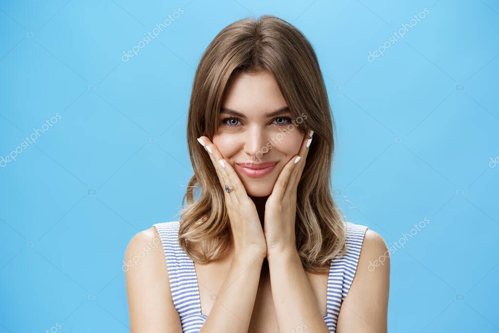Pretty european girl pressing palms to cheeks and smiling happily while looking at camera as if checking out appereance in mirror liking result after applying skincare treatment mask over blue wall. model released
