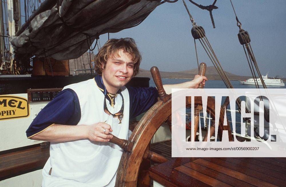 Tim Sander (Schauspieler) bei der Camel Trophy (Kapverdische Inseln) 03 00 mapo Mann geboren am 27.07.1978 in Berlin Sprecher Synchronsprecher Synchronsprechen Synchronisation Synchron Schauspiel TV Fernsehen Serie GZSZ Gute Zeiten, schlechte Zeiten RTL Rolle des Kai Scholl dunkelhaarig kurzhaarig Bart Bärtchen Ziegenbart Schmuck Ring leger T-Shirt bunt stehend draussen auf Boot Segelboot Schiff Segelschiff Ruder Steuerrad haltend festhaltend halb quer posierend lächelnd Wasser Meer See Ozean Horizont Fototermin Camel Trophy im März 2000 auf den Kapverdischen Inseln