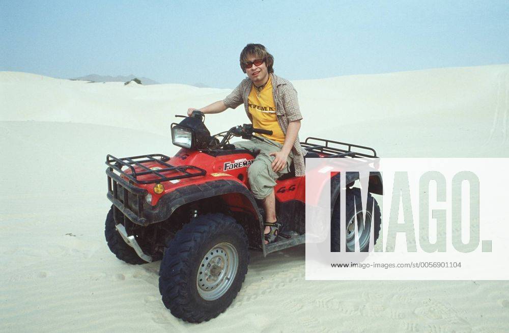 Tim Sander (Schauspieler) bei der Camel Trophy (Kapverdische Inseln) 03 00 mapo Mann Sprecher Synchronsprecher Synchronisation Schauspiel TV Fernsehen Serie GZSZ Gute Zeiten, schlechte Zeiten RTL Kai Scholl dunkelhaarig kurzhaarig Bart Bärtchen Ziegenbart Schmuck Kette Anhänger Ring Sonnenbrille leger Hemd kariert kleinkariert T-Shirt gelb Schrift Aufschrift Aufdruck One Life. Live it. Camel Trophy-Logo Hose Shorts Bermudas beige khaki Sandalen sitzend auf Fahrzeug Strandfahrzeug Quad Motorrad vier Räder vierrädrig Lenker Steuer haltend ganz quer lächelnd Strand Sand Wüste Horizont Fototermin Camel Trophy im März 2000 auf den Kapverdischen Inseln