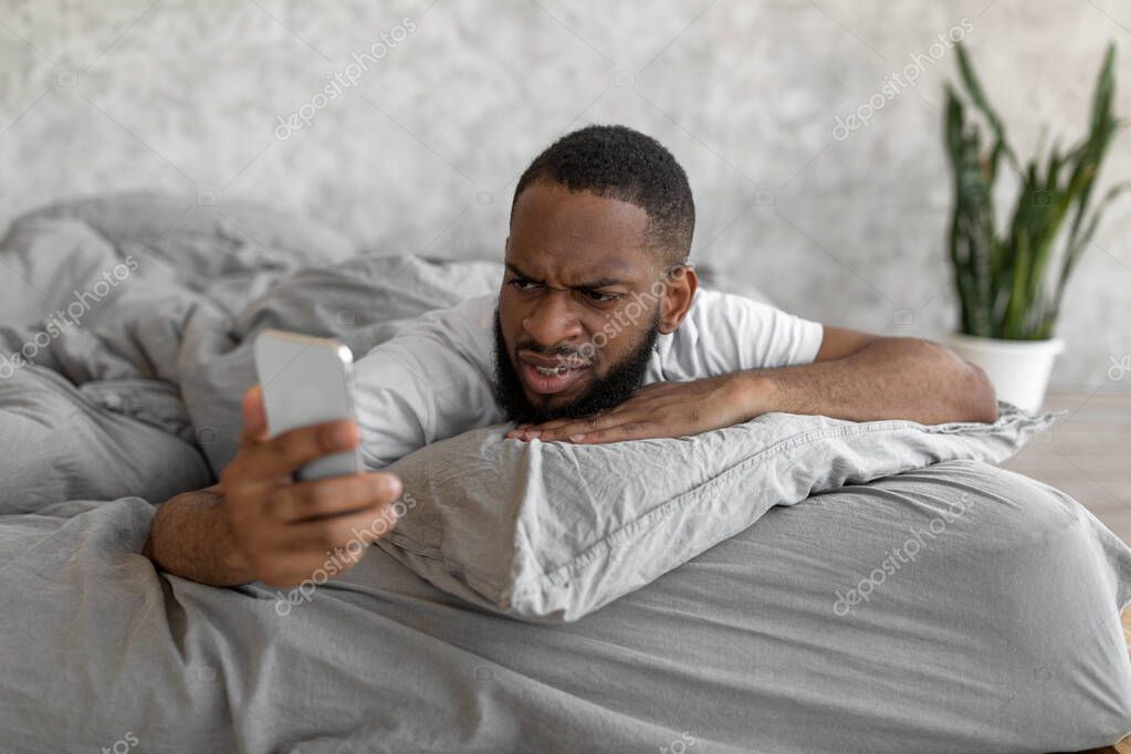 Oversleep Concept. Portrait of confused black man waking up with anxiety, upset angry young guy turned off alarm clock on smartphone lying on bed in bedroom, reading bad news, lookign at screen model released