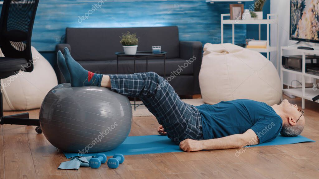 Retired person using fitness toning ball and lifting legs to stretch muscles. Aged man training and doing physical exercise while laying on yoga mat. Pensioner exercising with equipment model released