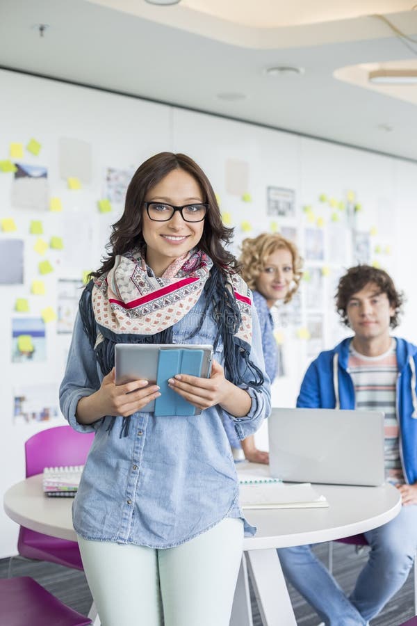 Portrait of smiling businesswoman holding digital tablet with colleagues in background at creative office, Portrait of smiling businesswoman holding digital tablet with colleagues in background at creative office
