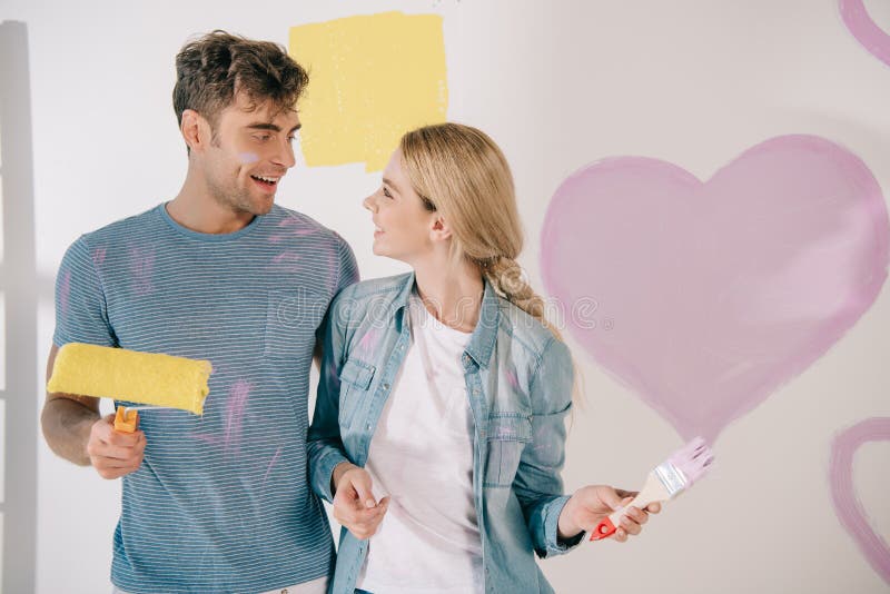 Young couple looking at each other while standing near pink heart painted on white wall, Happy young couple looking at each other while standing near pink heart painted on white wall