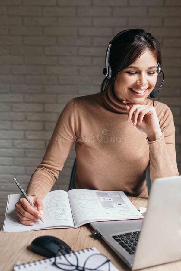 Smiling woman wearing headphones using laptop, writing notes, Smiling student woman wearing headphones using laptop, writing notes model released