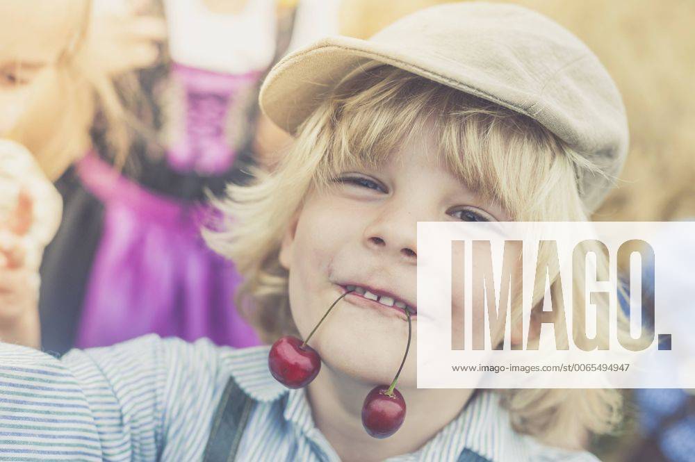 IMAGO Creative: Landleben - Essen Portrait of little boy holding two cherries with his mouth model released Symbolfoto MJF001587