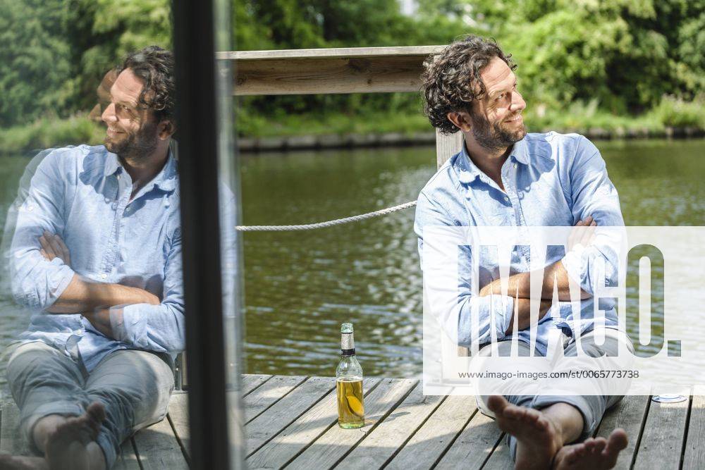 Smiling man sitting on platform at the waterside with beer bottle model released Symbolfoto property released FMKF001965