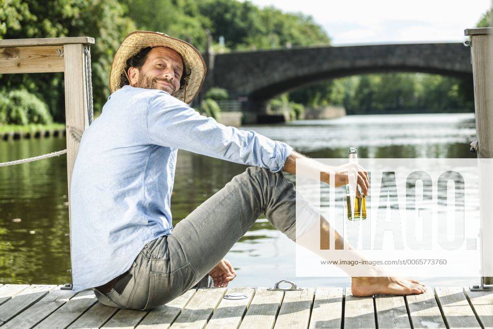 Smiling man sitting on platform at the waterside with beer bottle model released Symbolfoto property released FMKF001983
