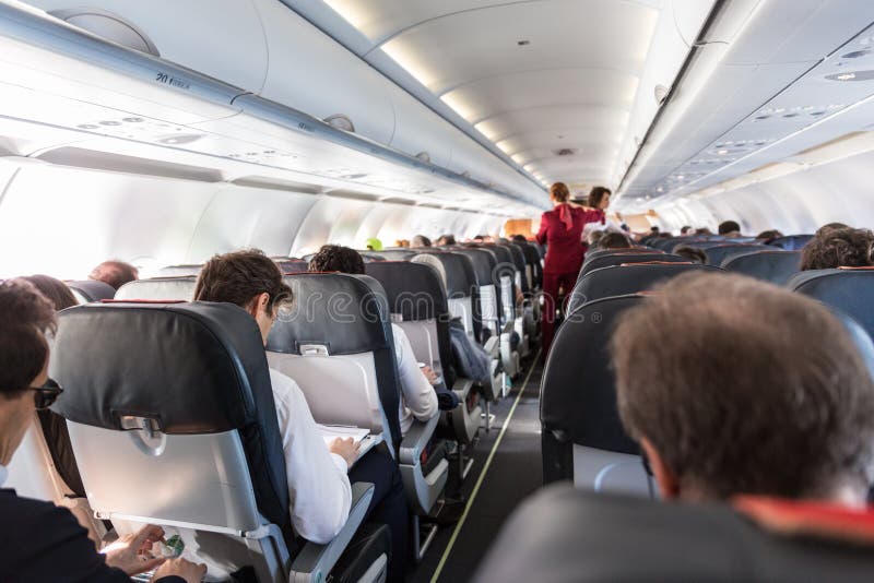 Interior of commercial airplane with passengers on their seats during flight., Interior of commercial airplane with unrecognizable passengers on their seats during flight. Stewardess in red uniform walking the aisle of commercial airplane.