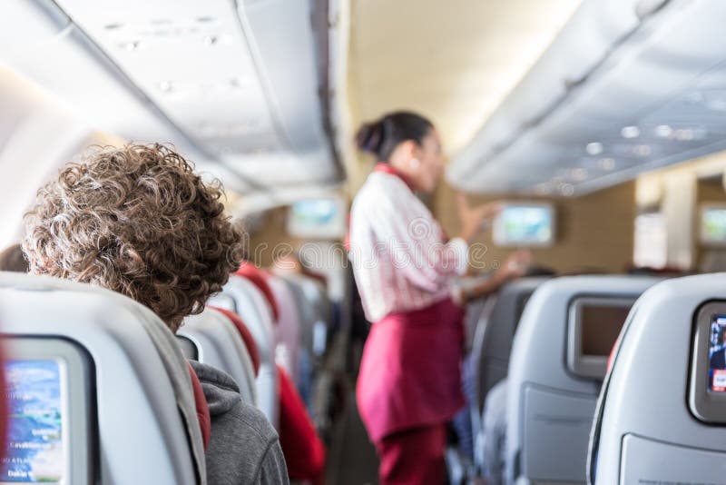 Interior of commercial airplane with passengers on their seats during flight., Interior of commercial airplane with unrecognizable passengers on their seats during flight. Stewardess in red uniform at the aisle of commercial airplane.