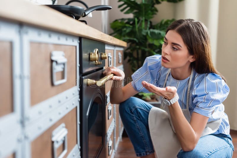 Young adult woman in apron looking, young adult woman in apron looking at timer on oven and gesturing with hand in modern kitchen ,stock image model released