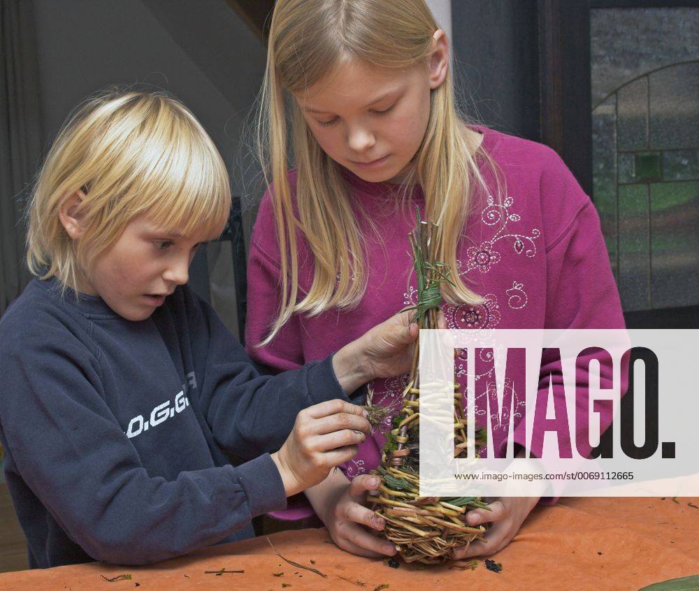 Zwei Kinder flechten aus Weidenzweigen eine Nistkugel fuer Voegel, Nest wird mit Moos und Schilfblueten ausgekleidet, Deutschland nesting material for ball nest, two children braiding willow branches and coating with moss and reeds, Germany BLWS416031