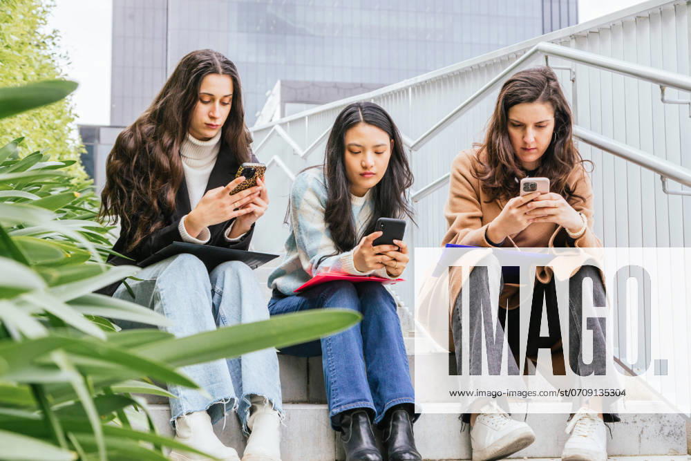 Three young female friends sit closely on outdoor stairs, each deeply engaged in her smartphone, surrounded by city buildings Model Released IgnacioFerrandiz_ID22609_659648_010