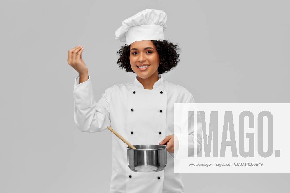 Culinary and people concept - happy smiling female chef in toque with saucepan cooking food showing gourmet sign over grey background. female chef with saucepan and spoon cooking food ,model released, Symbolfoto