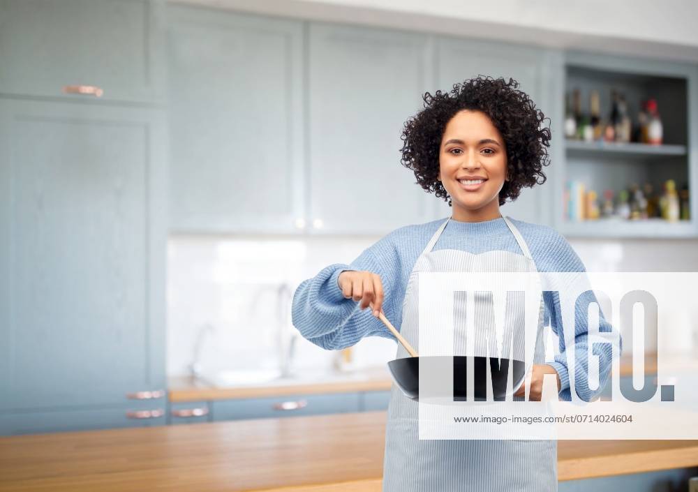 Culinary and people concept - happy smiling woman in apron with frying pan and spoon cooking food over home kitchen background. happy woman cooking food in frying pan in kitchen ,model released, Symbolfoto
