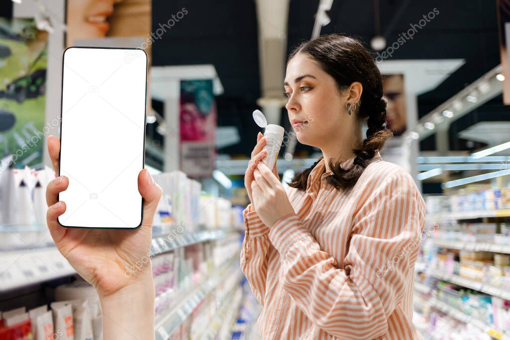 Portrait of Caucasian woman holds cream tube and sniffs fragrance of products. Choice of care cosmetics in supermarket. Hand shows smartphone with mock up. Concept of shopping and purchase.