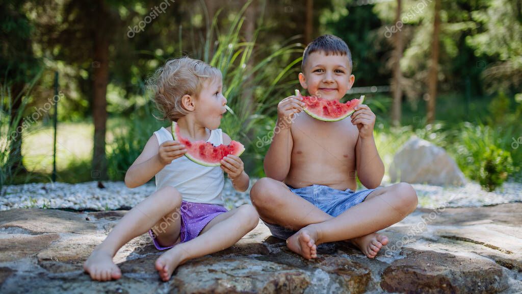 Little chidren sitting near a lake and eating watermelon on hot sunny day during summer vacation.