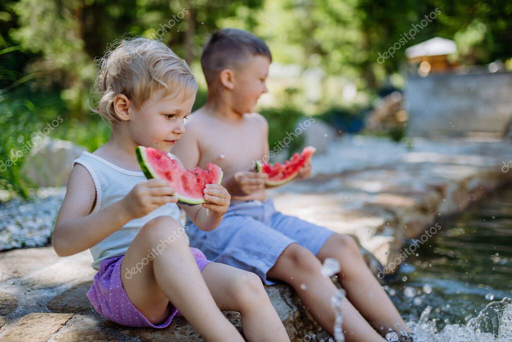 Little chidren sitting near a lake and eating watermelon on hot sunny day during summer vacation.
