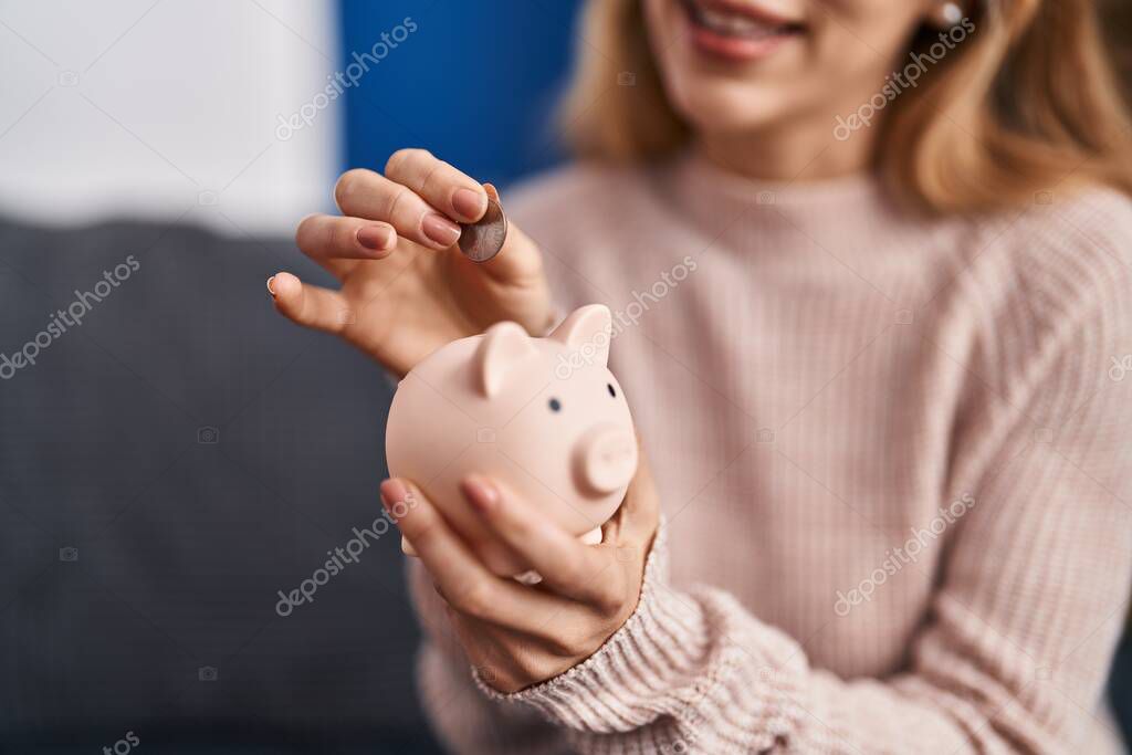 Young woman inserting coin on piggy bank at home