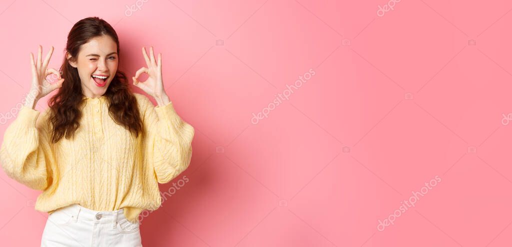 Portrait of cheerful pretty girl winking and saying yes, showing okay sign, alright gesture, praise good job, well done, standing pleased against pink background.