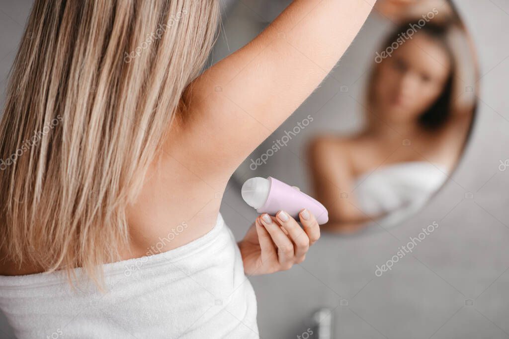 Young lady using antiperspirant stick for underarms for sweating prevention, woman applying deodorant to armpit after shower, standing in bathroom interior, selective focus