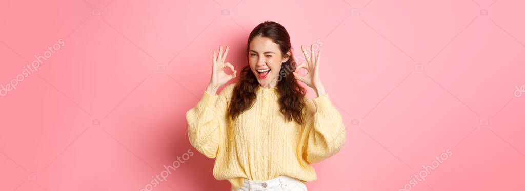 Portrait of cheerful pretty girl winking and saying yes, showing okay sign, alright gesture, praise good job, well done, standing pleased against pink background.