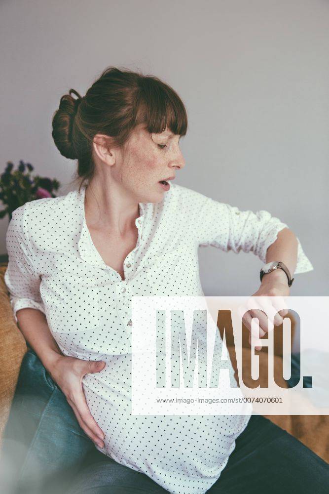 Expectant mother timing her contractions while sitting on couch at home model released Symbolfoto property released MFF03391
