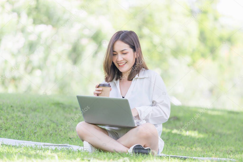 Young freelancer sitting with a laptop in a green park. Smiling modern woman using laptop and drinking takeaway coffee