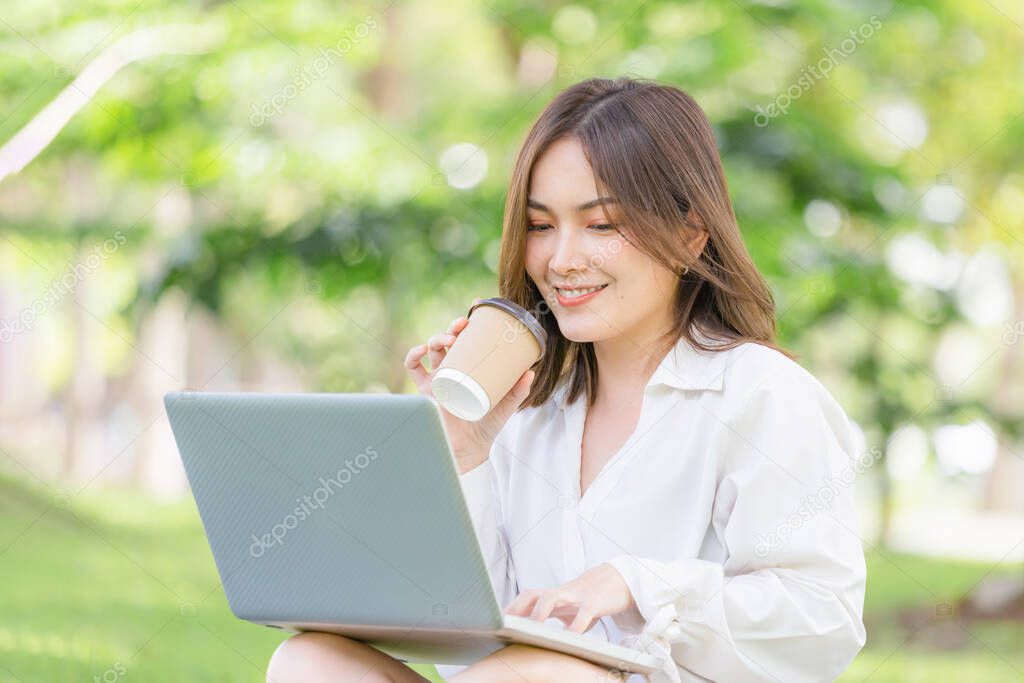 Young freelancer sitting with a laptop in a green park. Smiling modern woman using laptop and drinking takeaway coffee