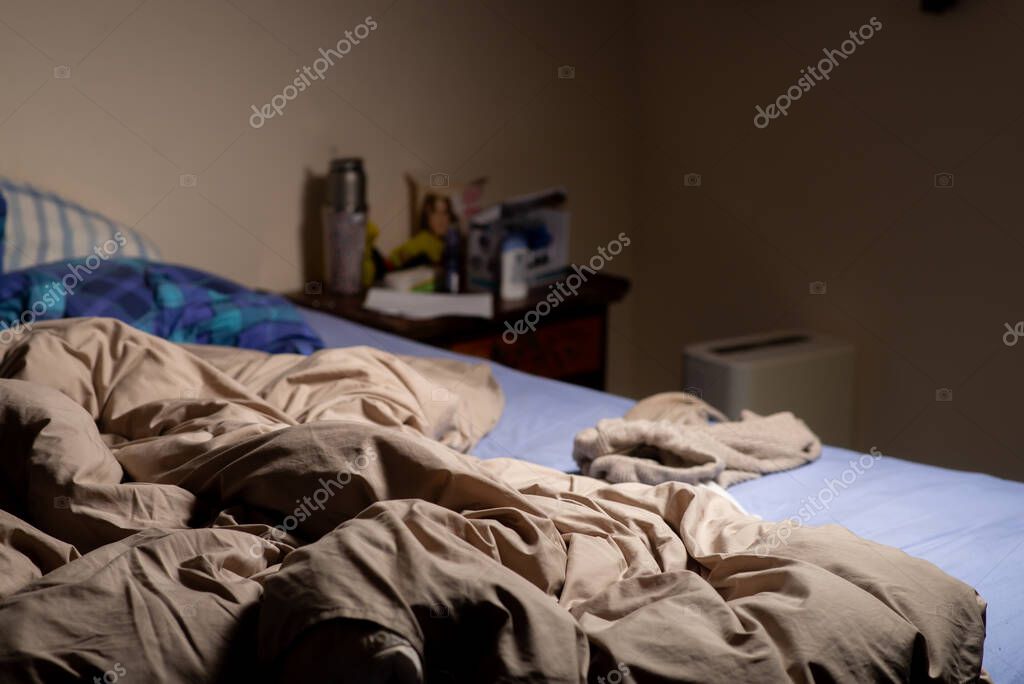 An empty unmade bed in the dark. A crumpled blanket lit by a lamp.