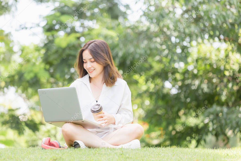 Young freelancer sitting with a laptop in a green park. Cheerful modern woman sitting in park while drinking takeaway coffee and look at laptop