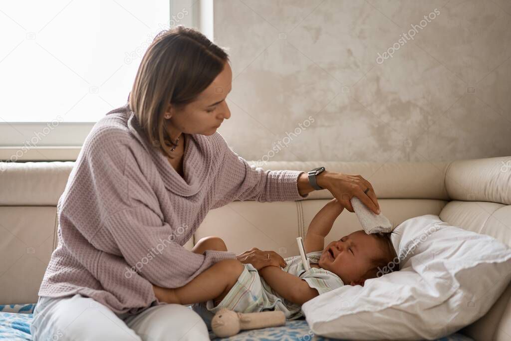 Worried young mother sitting on sofa beside her sick son with high fever. Mom measures temperature using thermometer of sick child lying under blanket at home.