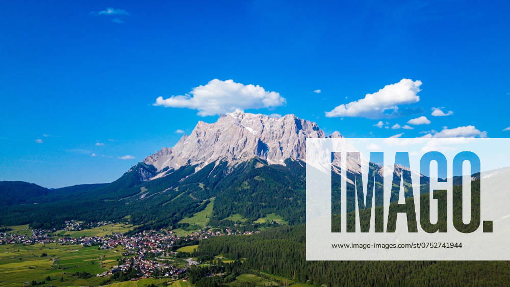 Zugspitze, höchster Berg Deutschlands, markante Felsformation, aufgenommen aus der Tiroler Zugspitz Arena, umgeben von dichten Wäldern und grünen Tälern, malerische Dörfer im Vordergrund, klarer blauer Himmel, weiße Wolken, majestätische Präsenz, beeindruckende Gipfelhöhe, beliebtes Ziel für Wanderer und Naturliebhaber, atemberaubende Alpenkulisse, harmonisches Zusammenspiel von Natur und Mensch, Sommerlandschaft, kontrastreiche Farben.