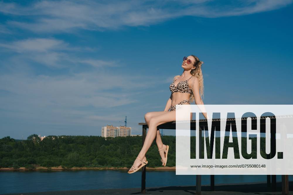 Woman in Leopard Swimsuit on Platform Over Water A woman in a leopard print swimsuit sits on a platform overlooking a body of water, with a clear blue sky and fluffy clouds in the background. She is wearing sunglasses and smiling at the camera. ,model released, Symbolfoto