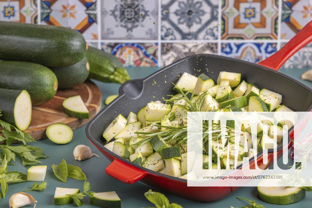 Diced courgettes in a red pan, flying herbs, stacked courgettes in the background in front of colourful tiles