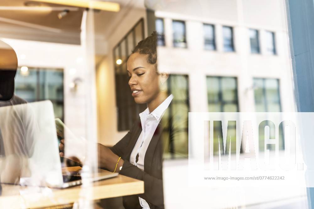 Blick von außen auf eine lateinamerikanische Geschäftsfrau, die in einer Cafeteria mit ihrem Smartphone arbeitet Views from outside the window of a latin businesswoman using smartphone working in a cafeteria