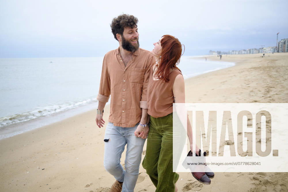 A joyful couple walks barefoot on a sandy beach, smiling and looking at each other with the sea in the background, Belgium bola05005