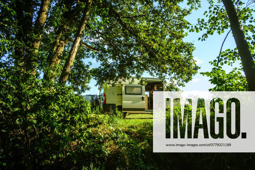 EIne Frau steht in der Schiebetür eines Campers, der in idyllischer Umgebung im Sommer auf einer Wiese zwischen Bäumen steht, Vogesen, Frankreich. Mit einem Campervan unterwegs